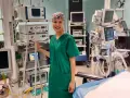 Uśmiechnięta pielęgniarka operacyjna, ubrana w zielony fartuch i czepkek, stoi obok stosu aparatury medycznej. Obok niej widać instrumentariuszka przygotowująca się do zabiegu.