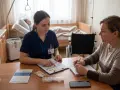 Podczas kursu opieki paliatywnej pielęgniarka omawia z pacjentką plan leczenia. W tle łóżko i wózek inwalidzki.
