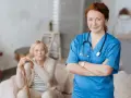Uśmiechnięta pielęgniarka w niebieskim uniformie, z pewnością ukończyła szybki kurs opiekuna medycznego. W tle starsza pani z laską.