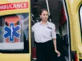Młoda kobieta w białej koszuli z naszywką "PARAMEDICS" stoi przy otwartych drzwiach karetki. Czy kierowca karetki musi być ratownikiem?