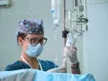 Pielęgniarka oddziałowa w czepku w gwiazdki, z maską i okularami, przygotowuje kroplówkę. Skrupulatność i troska to jej cechy.
