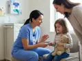 Pielęgniarka pediatryczna z uśmiechem pokazuje pulsoksymetr dziecku, które trzyma misia. Obok stoi mama.