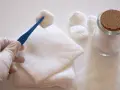 Rękawiczka w białej rękawiczce trzyma wacik na niebieskiej pęsecie, obok stosu gazy na ranę i słoika z wacikami.