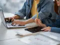 Dwie osoby pracują przy biurku, jedna pisze na laptopie, druga trzyma ołówek. Obok leży tablet i kalkulator. Może to być praca za najniższą krajową za godzinę.