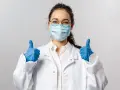 Uśmiechnięta pielęgniarka epidemiologiczna w masce i rękawiczkach pokazuje kciuki w górę.