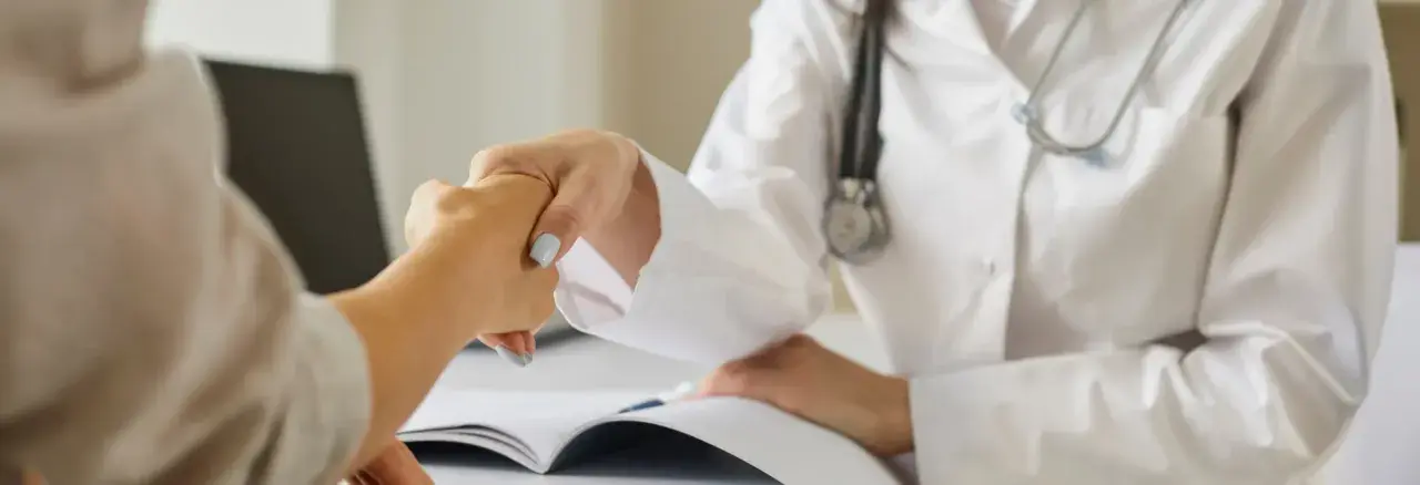 Lekarz i pacjent wymieniają uścisk dłoni podczas wywiadu. Na stole leży otwarta książka.