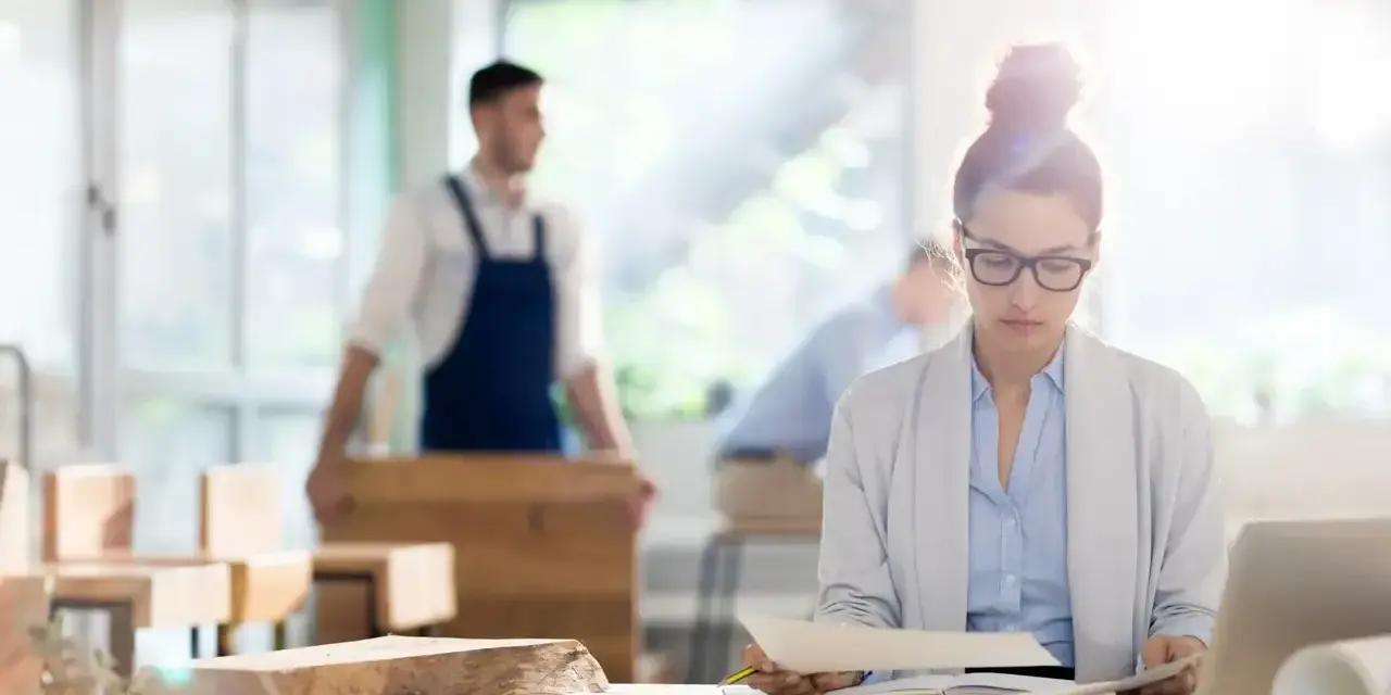 Kobieta w okularach analizuje dokumenty przy biurku, podczas gdy w tle mężczyzna w fartuchu pracuje przy drewnie. To jej staż z urzędu pracy.