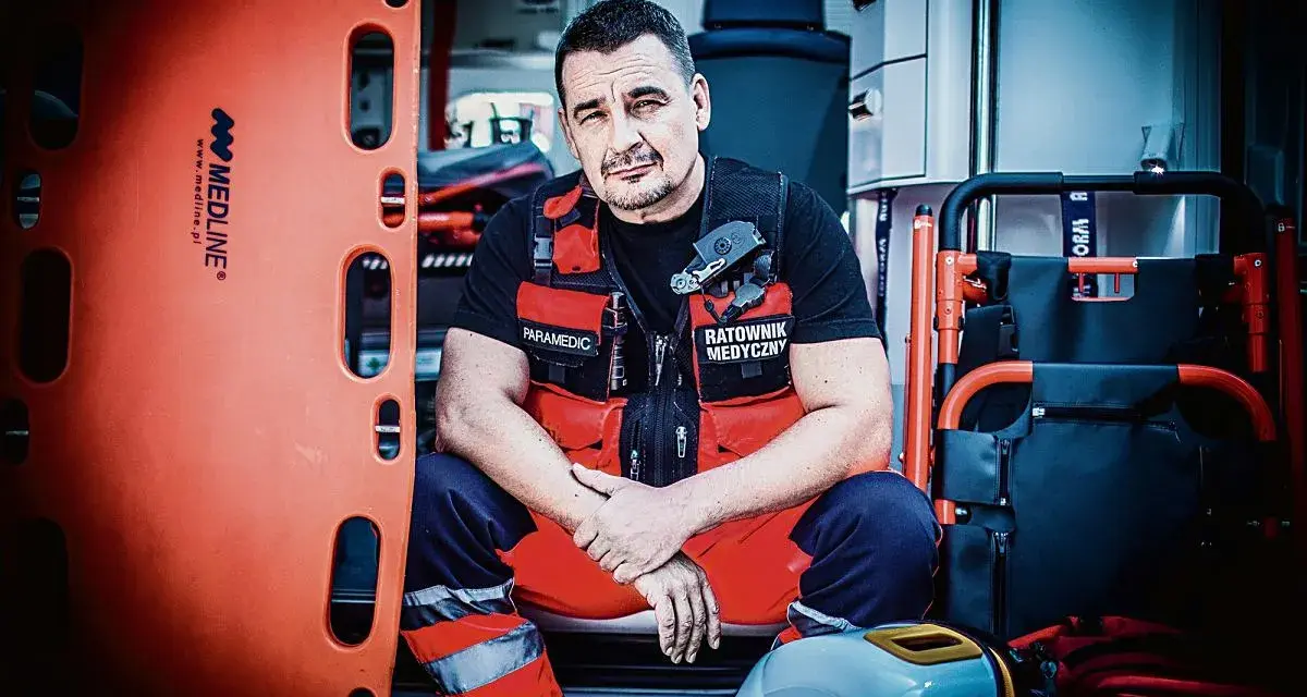 Ratownik medyczny w kamizelce z napisem "Paramedic" i "Ratownik Medyczny" siedzi w karetce obok noszy.