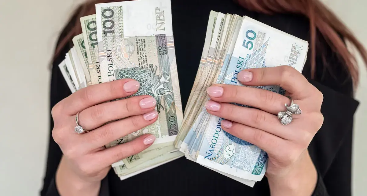 Dłonie z paznokciami w pastelowym różu trzymają pliki polskich banknotów 100 i 50 zł. To może być nagroda jubileuszowa.