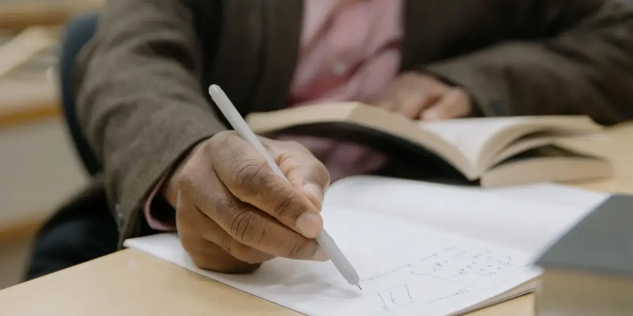 Osoba pisze ręcznie notatki w zeszycie, przygotowując list motywacyjny erasmus. W tle widać otwartą książkę.