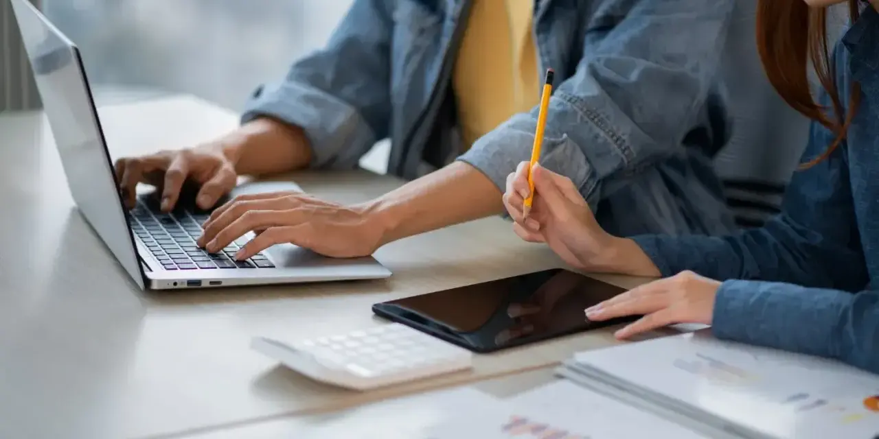 Dwie osoby pracują przy biurku, jedna pisze na laptopie, druga trzyma ołówek. Obok leży tablet i kalkulator. Może to być praca za najniższą krajową za godzinę.