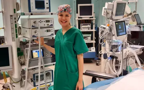Uśmiechnięta pielęgniarka operacyjna, ubrana w zielony fartuch i czepkek, stoi obok stosu aparatury medycznej. Obok niej widać instrumentariuszka przygotowująca się do zabiegu.