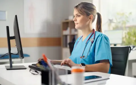 Pielęgniarka w niebieskim uniformie pracuje przy komputerze. Jej specjalizacja pielęgniarska to 5 lat doświadczenia.