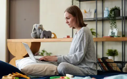 Kobieta w piżamie pisze na laptopie, przygotowując wniosek o urlop macierzyński. Obok pluszowy królik i zabawki.