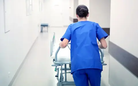 Salowa w niebieskim uniformie pcha łóżko szpitalne korytarzem. To część jej obowiązków w szpitalu.