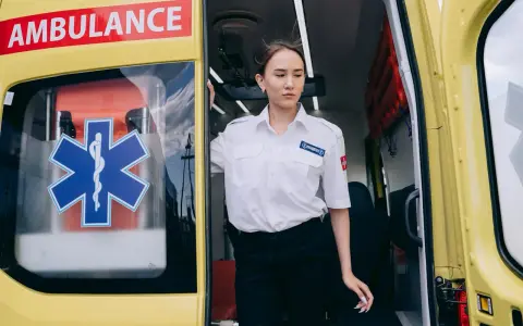 Młoda kobieta w białej koszuli z naszywką "PARAMEDICS" stoi przy otwartych drzwiach karetki. Czy kierowca karetki musi być ratownikiem?