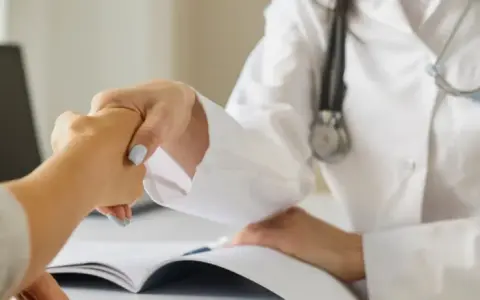 Lekarz i pacjent wymieniają uścisk dłoni podczas wywiadu. Na stole leży otwarta książka.