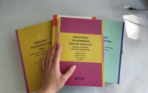 Ręka z pierścionkiem przegląda kolorowe czasopisma pielęgniarskie: "Podstawy Pielęgniarstwa", "Umiejętności Pielęgniarskie" i "Wybrane Umiejętności".
