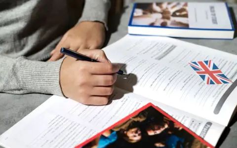 Osoba pisze długopisem w zeszycie z ćwiczeniami z języka angielskiego. Obok leży książka i flaga Wielkiej Brytanii. To może być przygotowanie do napisania klauzuli CV po angielsku.