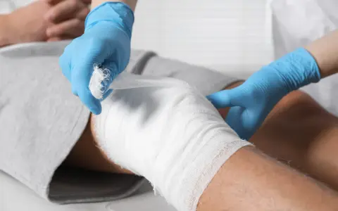 Ręce w niebieskich rękawiczkach opatrują kolano bandażem. Pokazane są różne sposoby opatrywania ran.