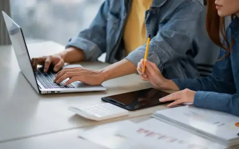 Dwie osoby pracują przy biurku, jedna pisze na laptopie, druga trzyma ołówek. Obok leży tablet i kalkulator. Może to być praca za najniższą krajową za godzinę.