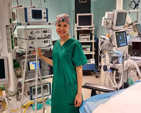 Uśmiechnięta pielęgniarka operacyjna, ubrana w zielony fartuch i czepkek, stoi obok stosu aparatury medycznej. Obok niej widać instrumentariuszka przygotowująca się do zabiegu.