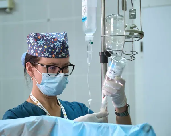 Pielęgniarka oddziałowa w czepku w gwiazdki, z maską i okularami, przygotowuje kroplówkę. Skrupulatność i troska to jej cechy.