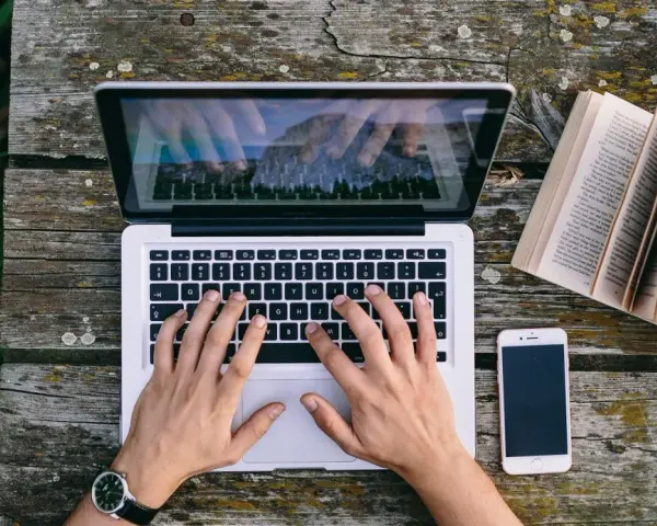 Ręce piszą CV funkcjonalne na laptopie, obok leży otwarta książka i telefon.