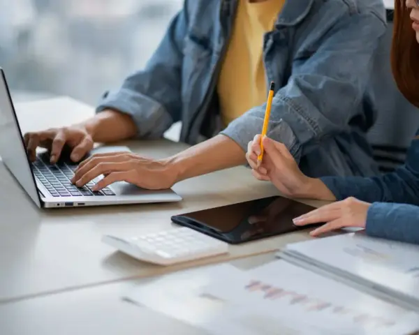 Dwie osoby pracują przy biurku, jedna pisze na laptopie, druga trzyma ołówek. Obok leży tablet i kalkulator. Może to być praca za najniższą krajową za godzinę.
