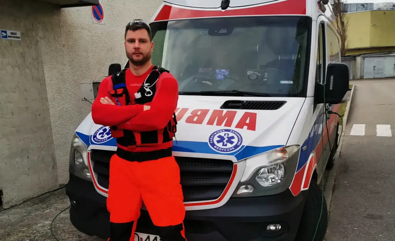 Ratownik medyczny w pomarańczowych spodniach i czerwonej bluzie stoi przed karetką. To jego praca, gotowy do akcji.