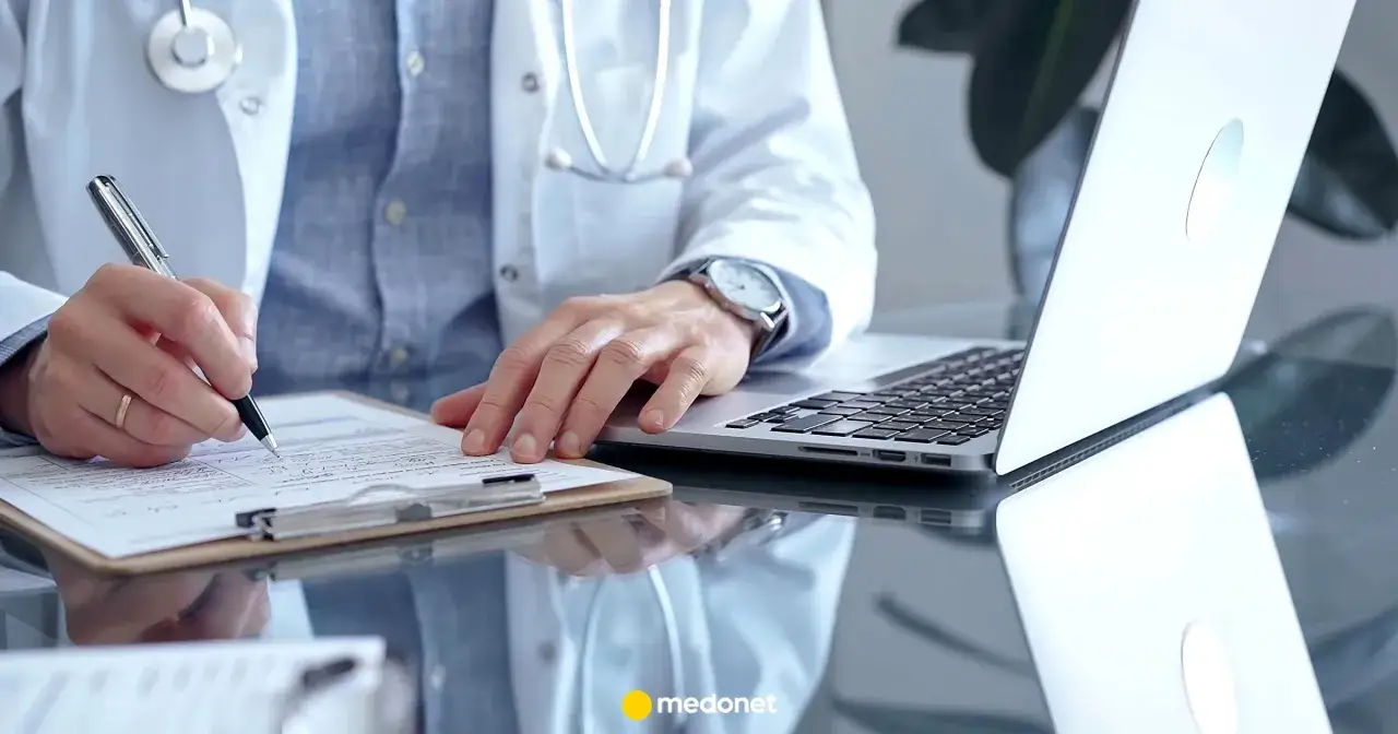 Lekarz w białym kitlu pisze na dokumentach, obok laptop. To może być zwolnienie lekarskie od psychiatry płatne 100%.
