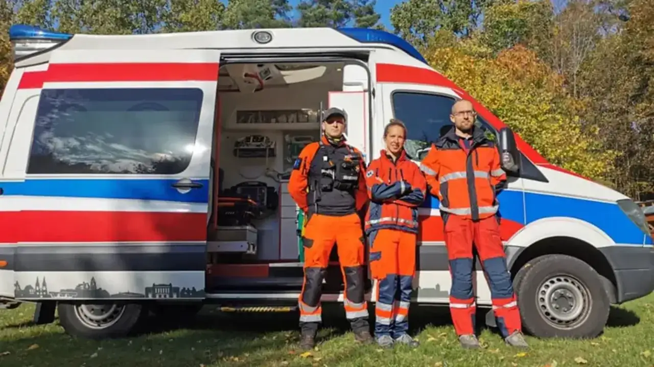 Trzech ratowników medycznych w pomarańczowych strojach stoi obok karetki. Zastanawiasz się, ile godzin pracuje ratownik medyczny?